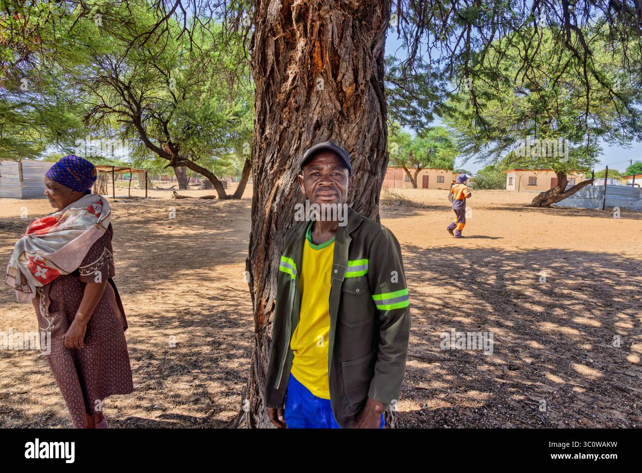 village, portrait d'un vieil homme souriant africain, à l'ombre d'un arbre, maisons en arrière-plan, personnes et passants Banque D'Images