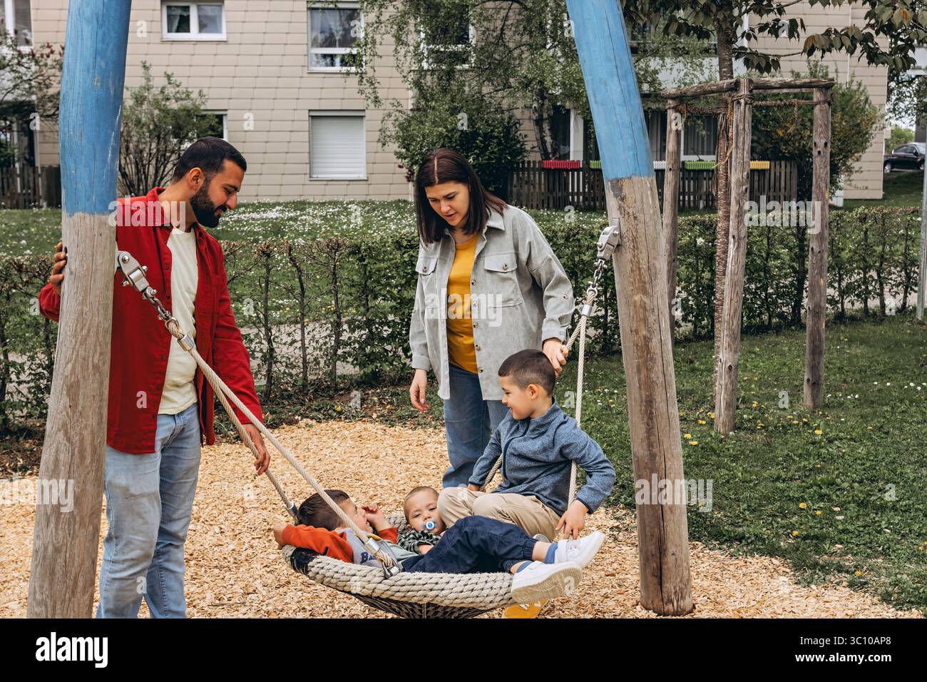 Famille heureuse avec deux fils jouant ensemble sur une grande balançoire de corde à l'aire de jeux. Les parents sourient, tissent des liens et passent du temps de qualité avec les enfants à l'extérieur Banque D'Images