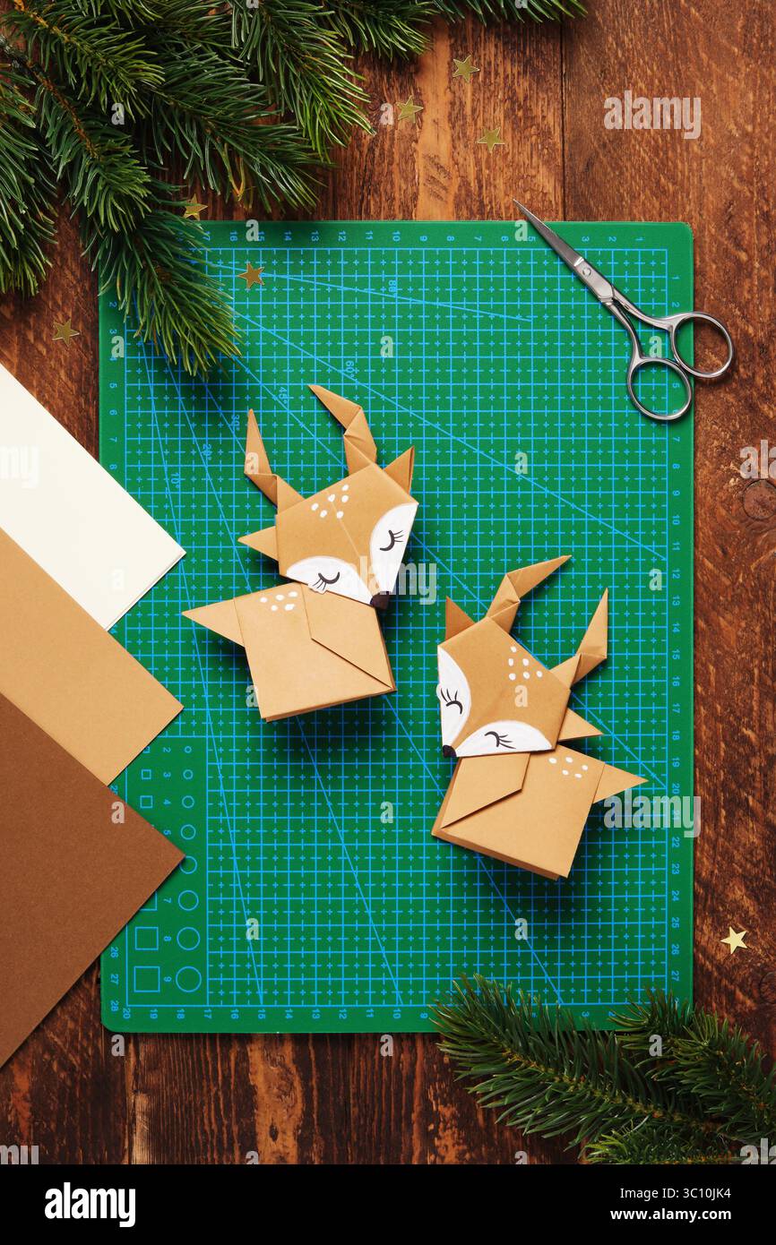 Deux cerfs en origami en papier brun sur un tapis de coupe vert. À proximité se trouvent des branches de sapin, du papier origami et des ciseaux. Banque D'Images
