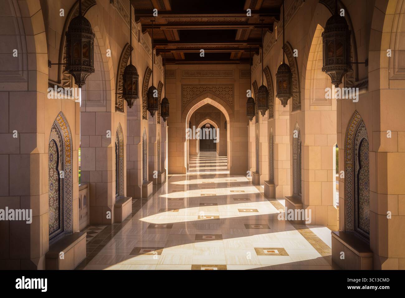 Vue sur un couloir baigné de soleil avec des arches ornées et des lanternes suspendues projetant des ombres sur le sol poli, Muscat, gouvernorat de Muscat, Oman. Banque D'Images