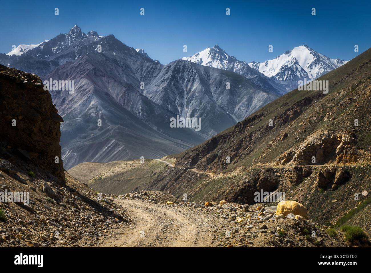 Vue sur un chemin de terre accidenté et sinueux serpentant à travers une vallée rocheuse et dure, menant vers des montagnes imposantes couvertes de neige brillante sous un ciel clair, le Tadjikistan. Banque D'Images