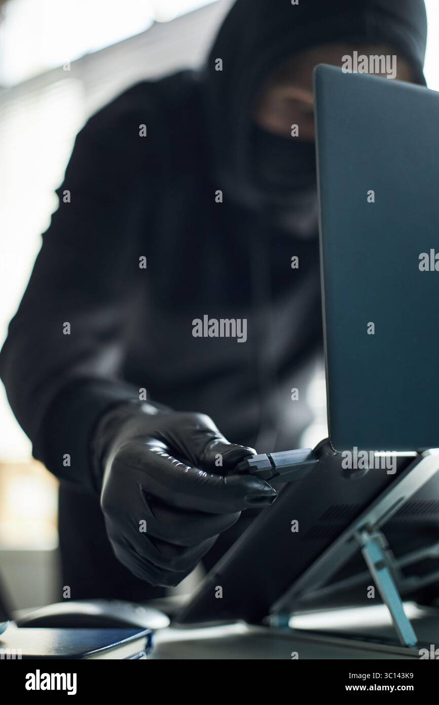 Homme méconnaissable portant des gants noirs et un sweat à capuche insérant un lecteur flash USB avec un logiciel malveillant dans un ordinateur portable pour pirater le système informatique de l'entreprise, se livrant à une activité criminelle secrète dans l'espace de travail Banque D'Images