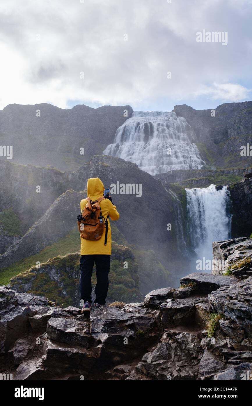 Un voyageur vêtu d'une veste de pluie jaune vif se tient sur un terrain rocheux, capturant les cascades frappantes qui cascadent dans le paysage sauvage islandais. Cascade de Dynjandi, Westfjord, Islande Banque D'Images