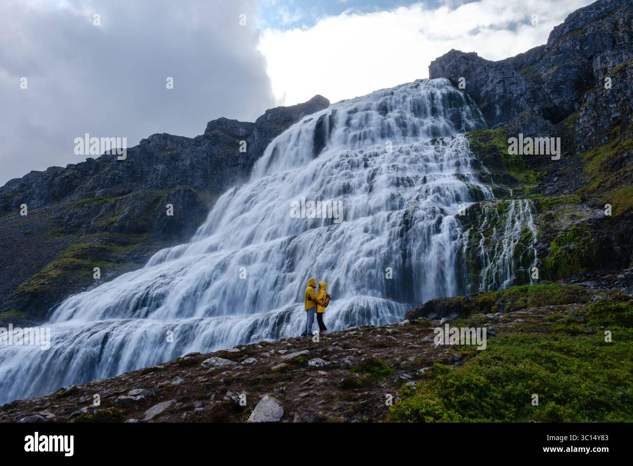 Une superbe cascade descend les falaises rocheuses d'Islande, avec un passionné de plein air vêtu d'une veste de pluie jaune éclatante debout en dessous, profitant de la vue à couper le souffle. Dyjandi Westfjords Islande Banque D'Images