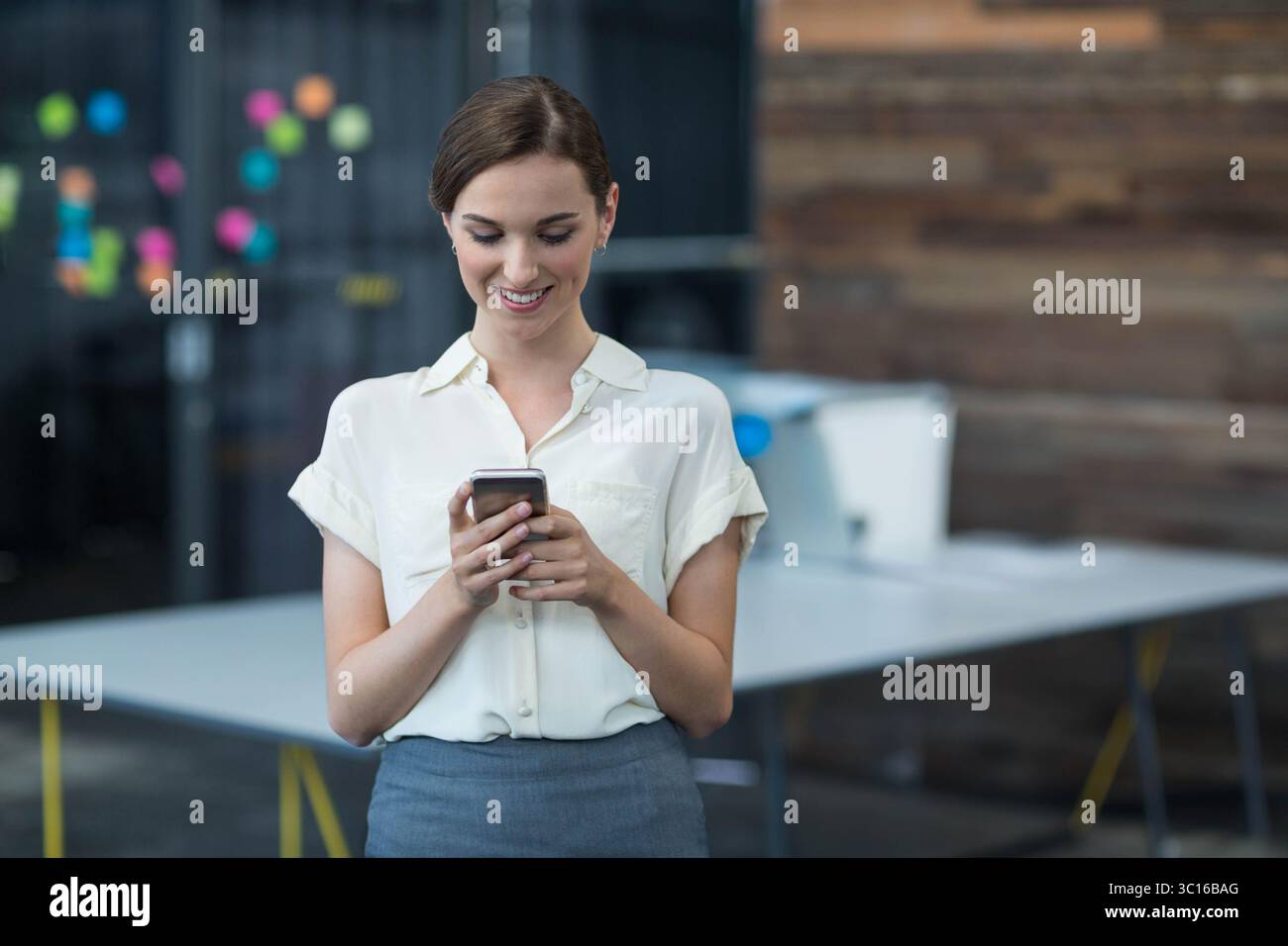 Employée de bureau adolescente vérifiant smartphone au bureau blanc avec imprimante, notes adhésives Banque D'Images