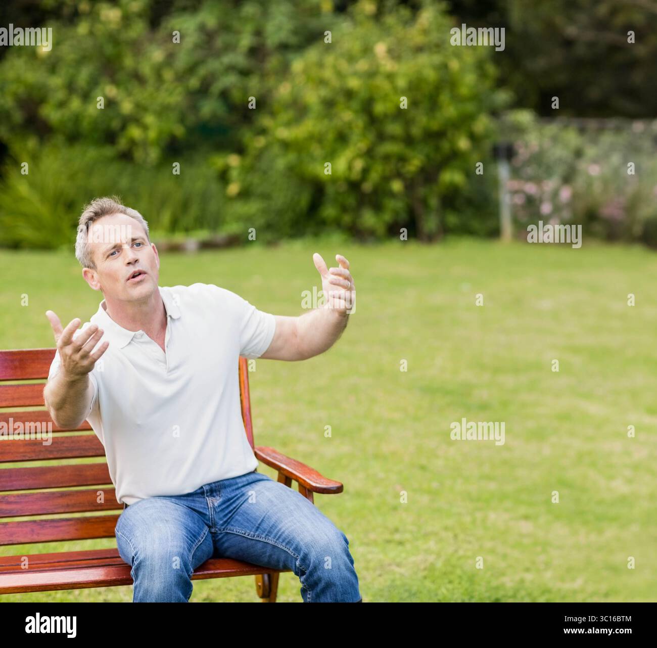 Homme assis sur un banc en bois sur une pelouse bien entretenue avec toile de fond de haie sous le ciel ensoleillé, espace de copie Banque D'Images
