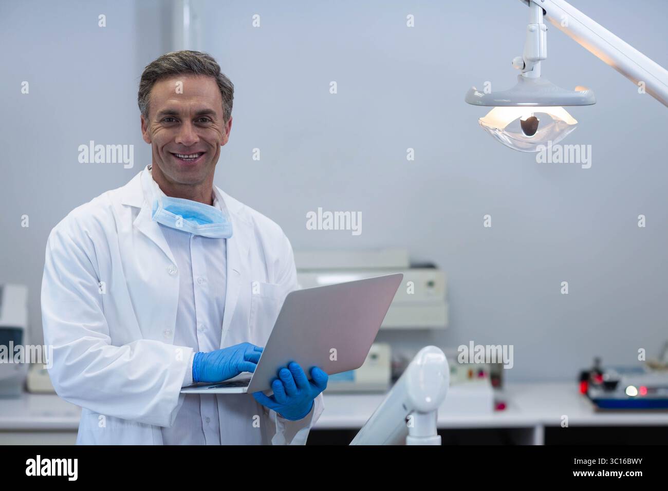 Dentiste masculin portant une blouse de laboratoire blanche tenant un ordinateur portable, souriant dans une clinique dentaire avec lampe, espace de copie Banque D'Images