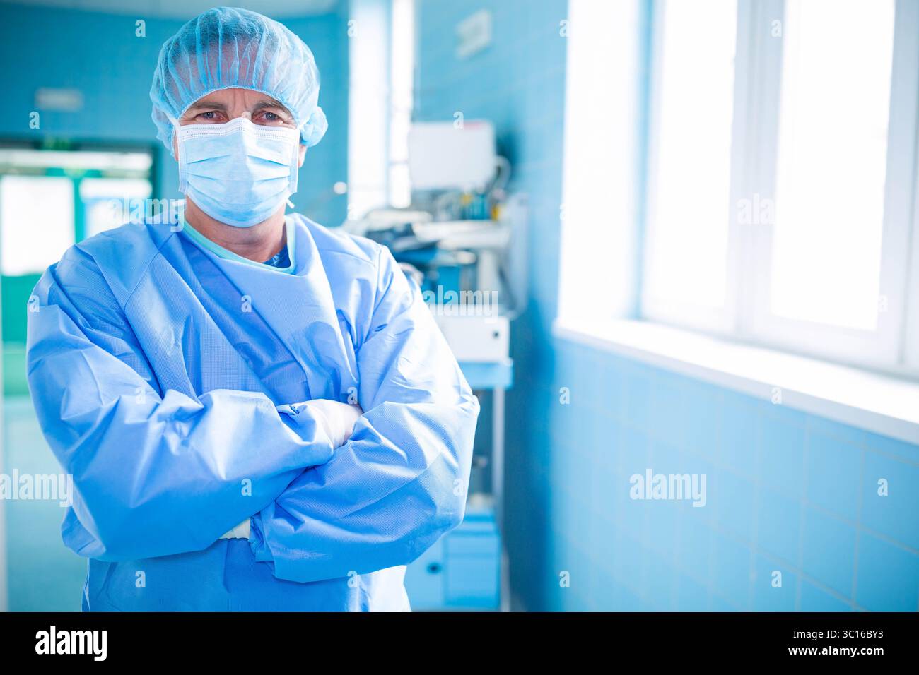Chirurgien masculin debout dans le couloir chirurgical carrelé bleu à côté du chariot d'équipement sous des fenêtres lumineuses Banque D'Images