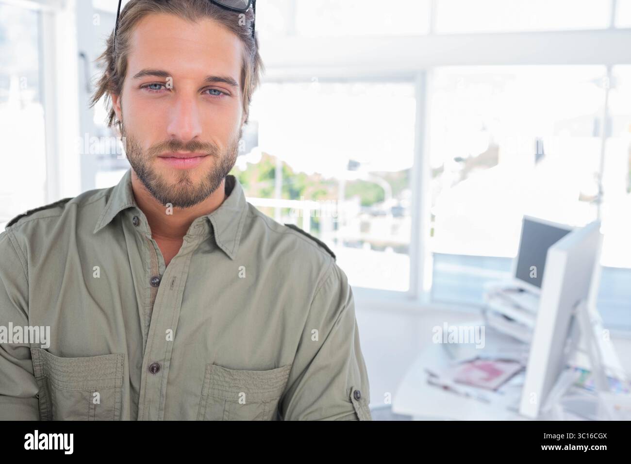Homme barbu debout près du bureau avec écran, papiers, lunettes de soleil et chemise vert olive Banque D'Images