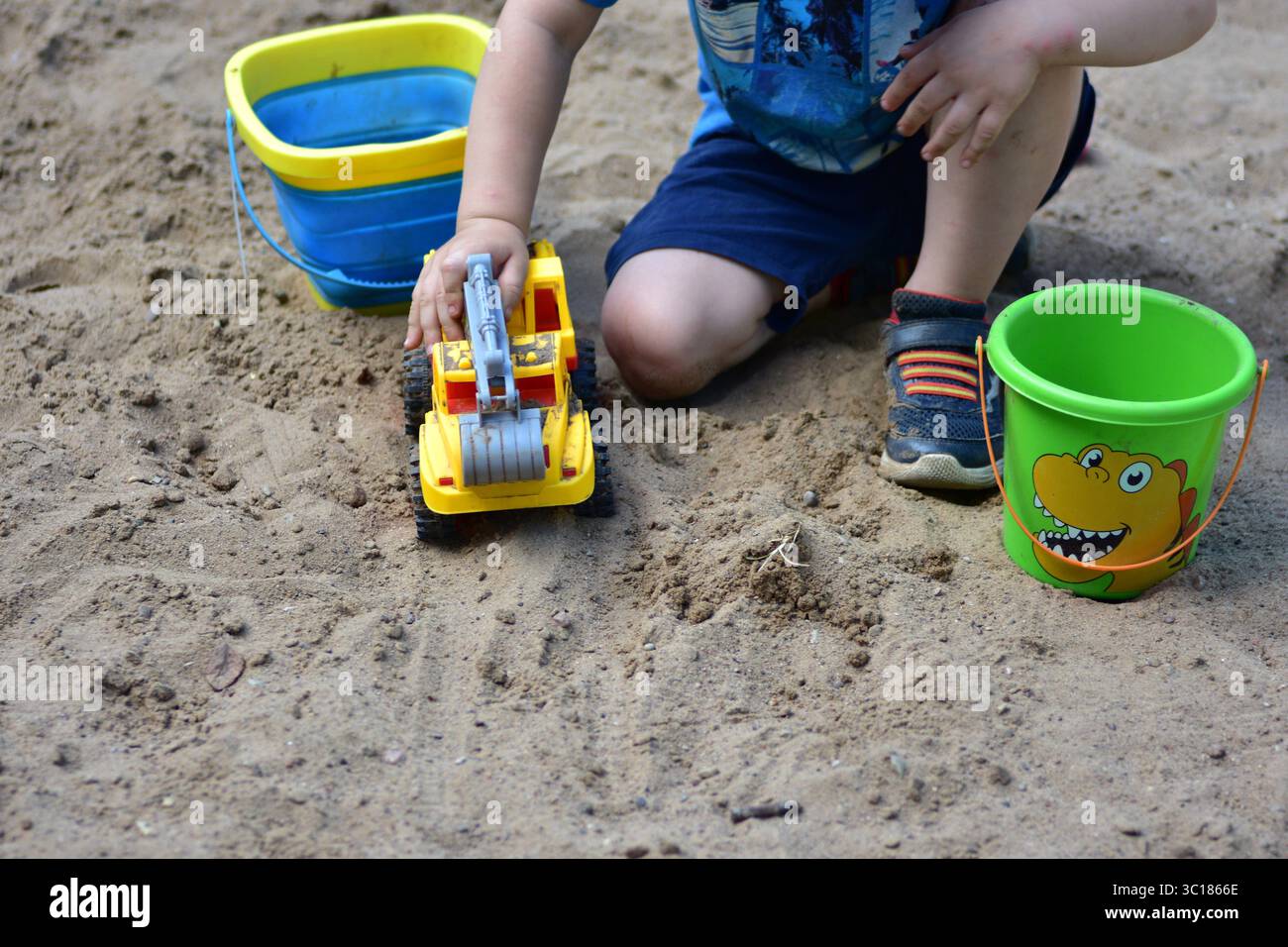 Garçon en bas âge anonyme jouant dans le bac à sable avec un camion-jouet jaune et deux seaux, entouré de sable sec par une journée ensoleillée, capturé au début du chi Banque D'Images