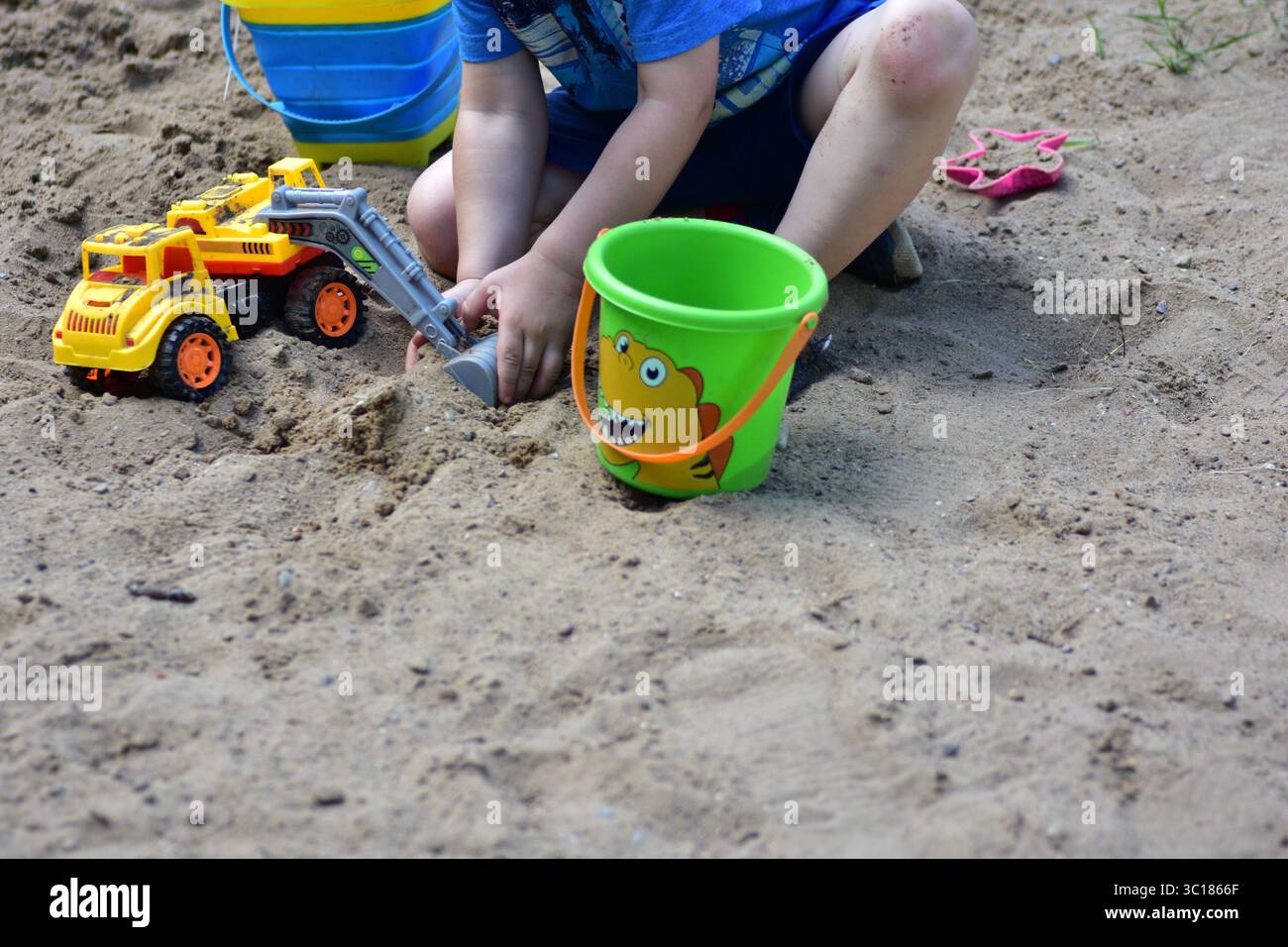 Garçon jouant dans le bac à sable avec un camion-jouet jaune et deux seaux, entouré de sable sec et d'herbe par une journée ensoleillée, capturé pendant la petite enfance p Banque D'Images