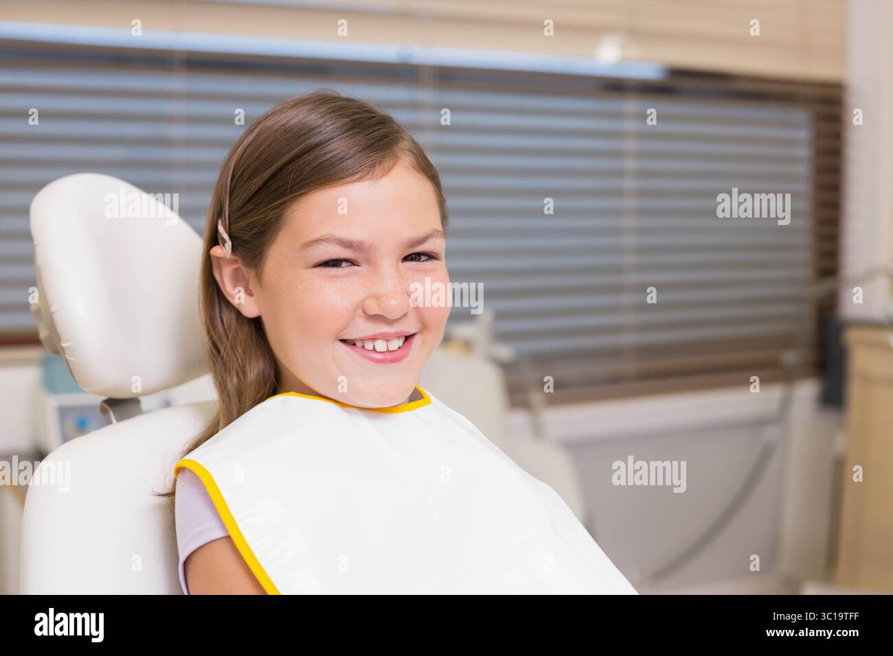 Patiente enfant de sexe féminin assise dans un fauteuil de clinique dentaire portant un bavoir souriant et attendant des soins Banque D'Images