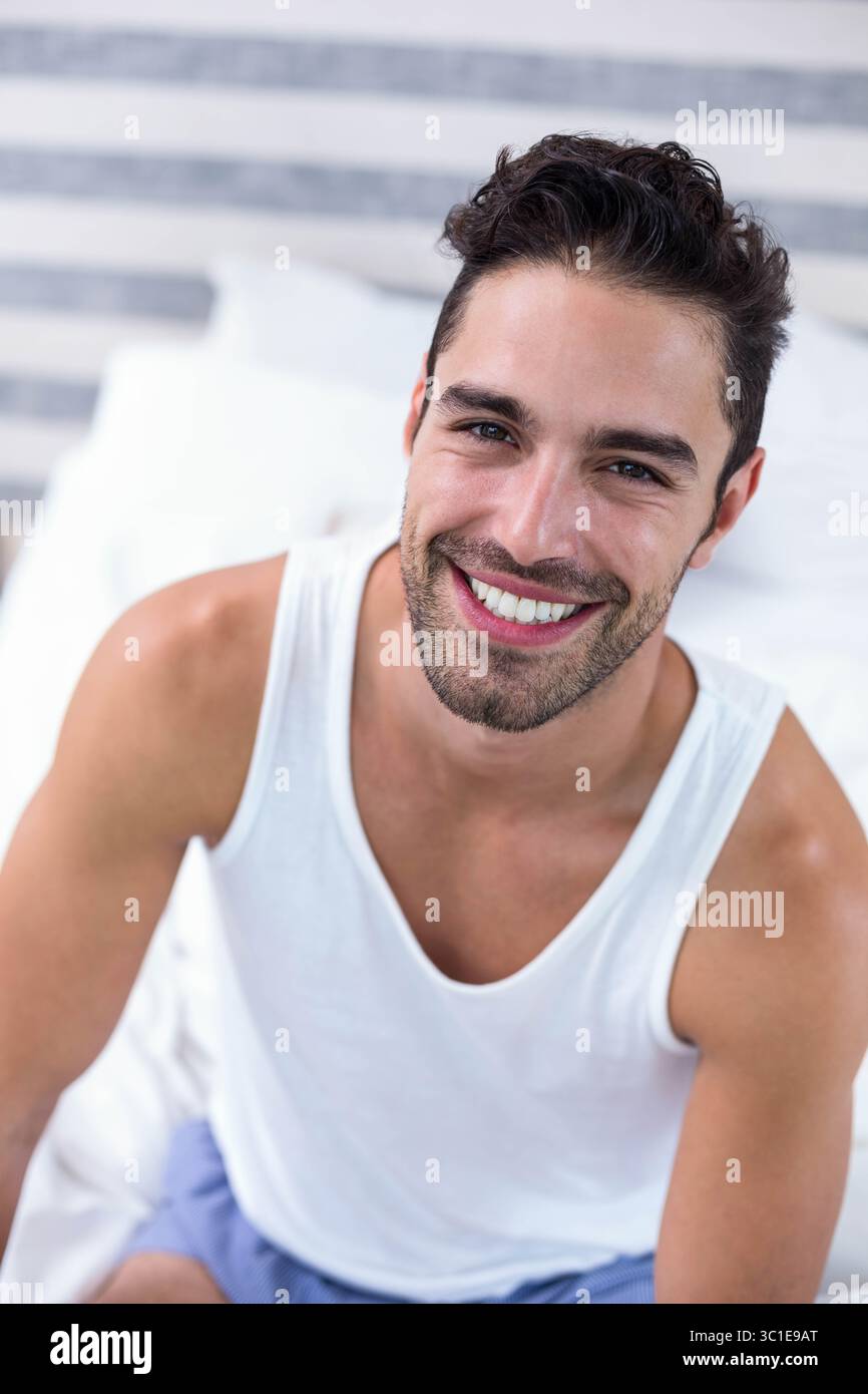 Homme assis sur le lit souriant vers le haut dans la chambre avec tête de lit rayée portant débardeur et short Banque D'Images