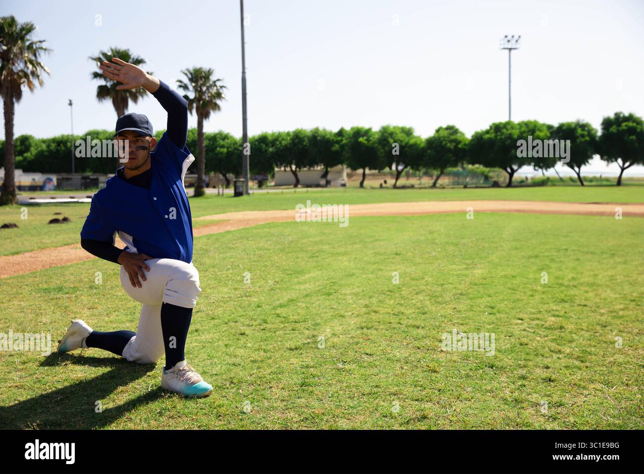 Adolescent garçon étirant le bras droit tout en s'agenouillant sur le terrain de baseball près de la terre à l'intérieur du champ, copier l'espace Banque D'Images