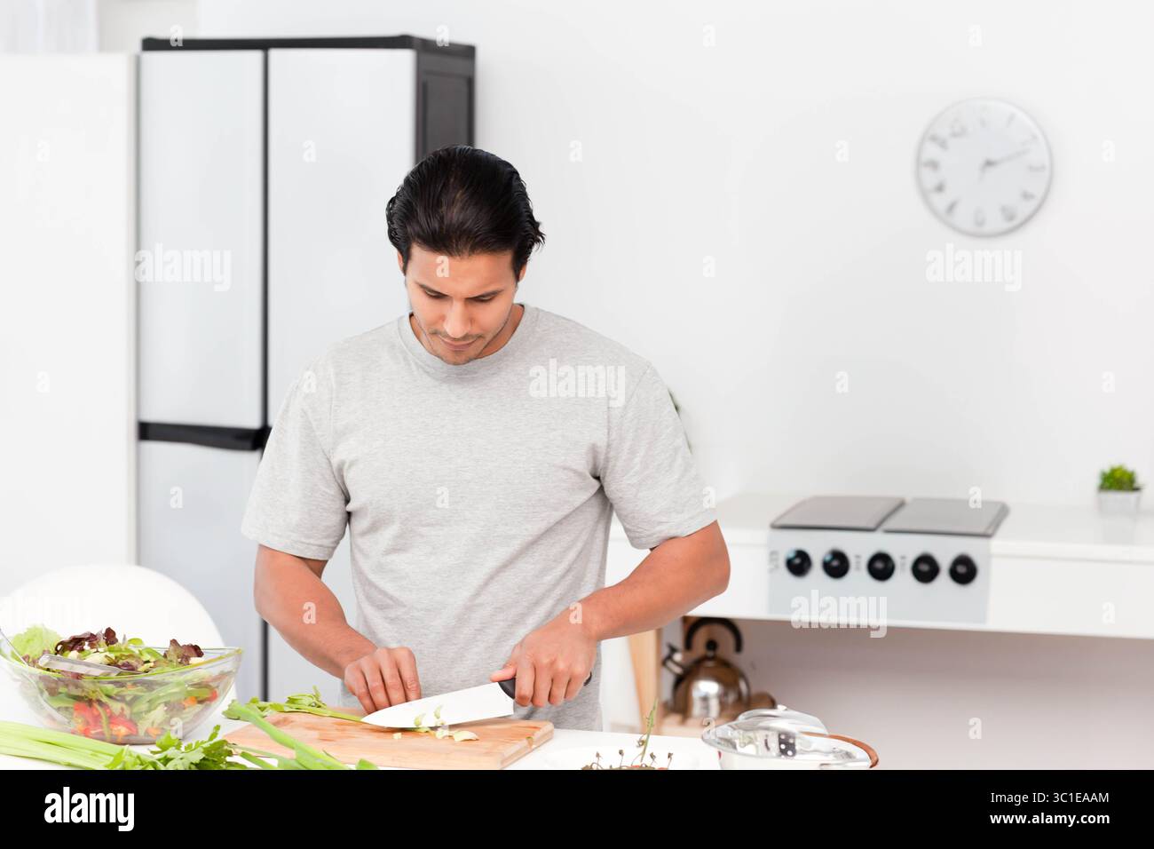 Mâle adulte moyen debout dans la cuisine blanche tranchant le céleri sur une planche à découper en bois à côté d'un bol en verre Banque D'Images