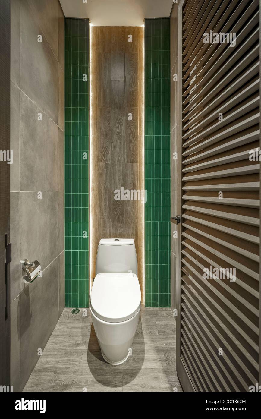 Cabine de toilette moderne avec carreaux verticaux verts éclatants, mur d'accent en bois et éclairage ambiant à bande LED. Toilettes publiques épurées et minimalistes. Banque D'Images