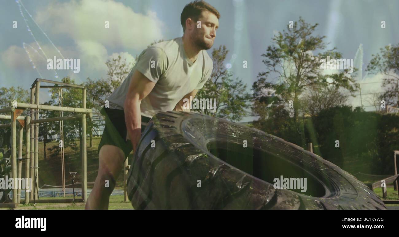 Homme de contrainte en chemise grise et short retournant le pneu de tracteur sur le parcours d'obstacle, avec des cadres en bois Banque D'Images