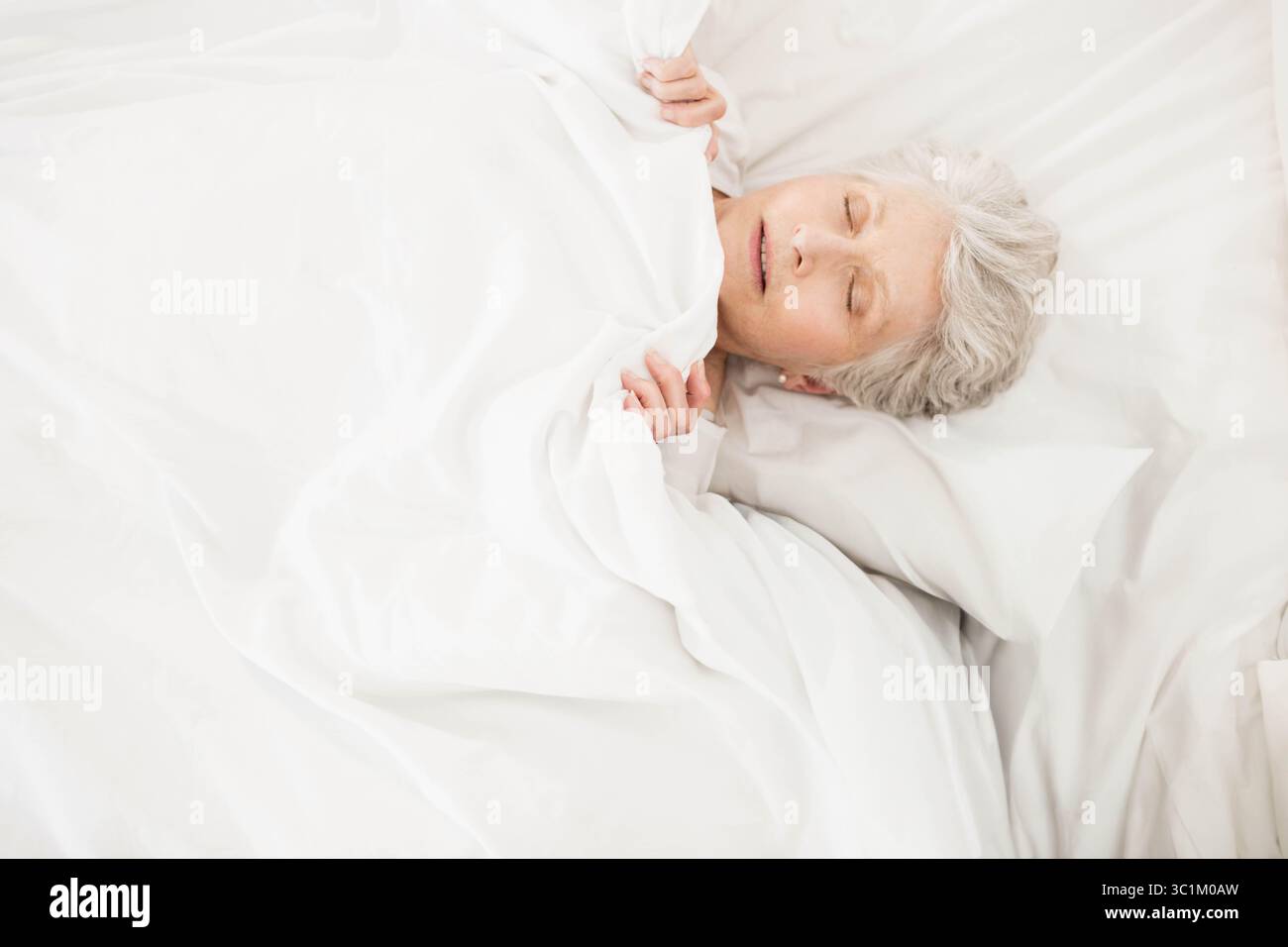 Femme senior dormant tranquillement dans la chambre à coucher à la maison sous des draps blancs et une couverture, espace de copie Banque D'Images