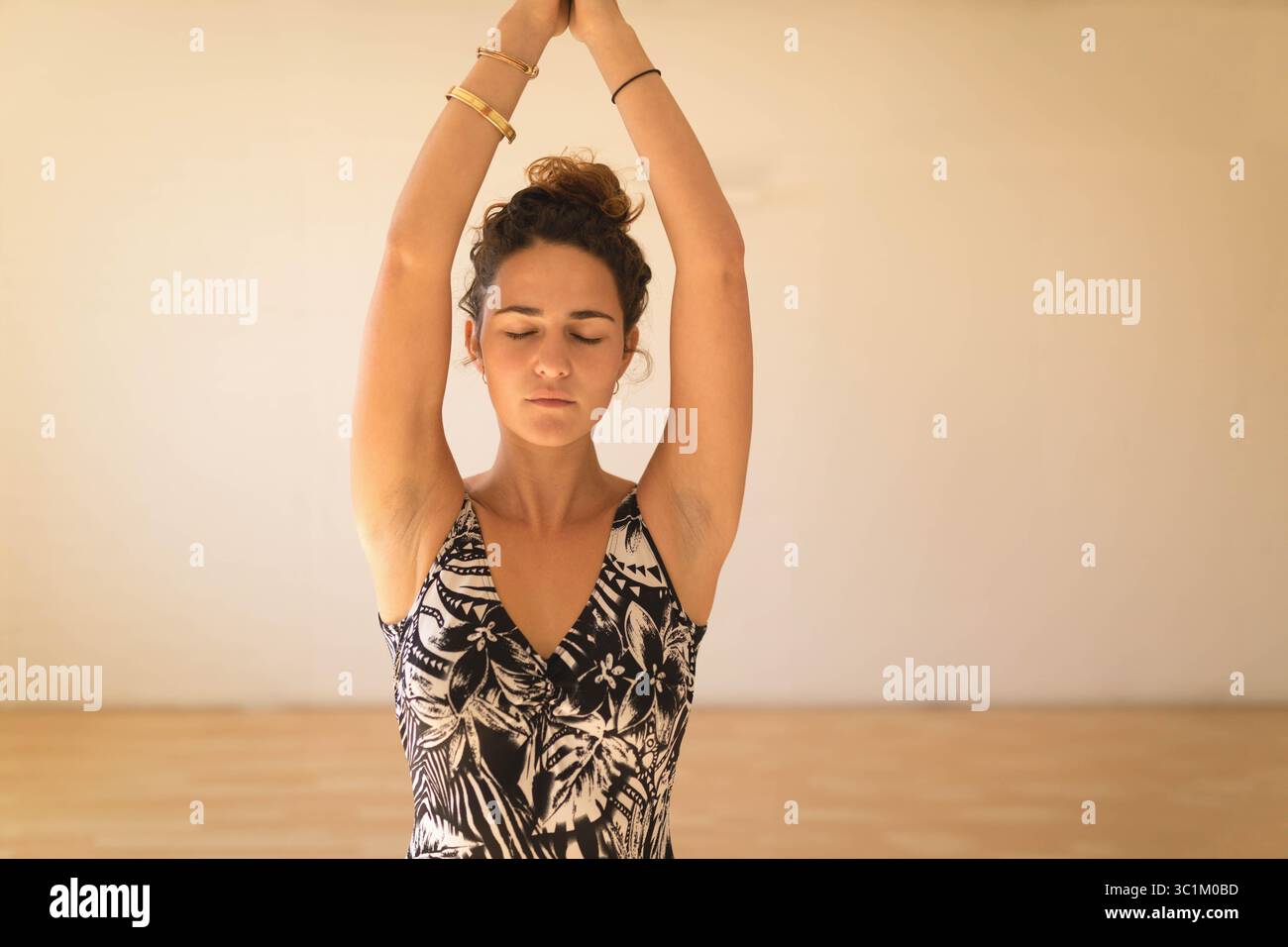 Femme adulte portant débardeur floral étirant les bras au-dessus de la tête dans l'espace de yoga à la maison avec des bracelets Banque D'Images