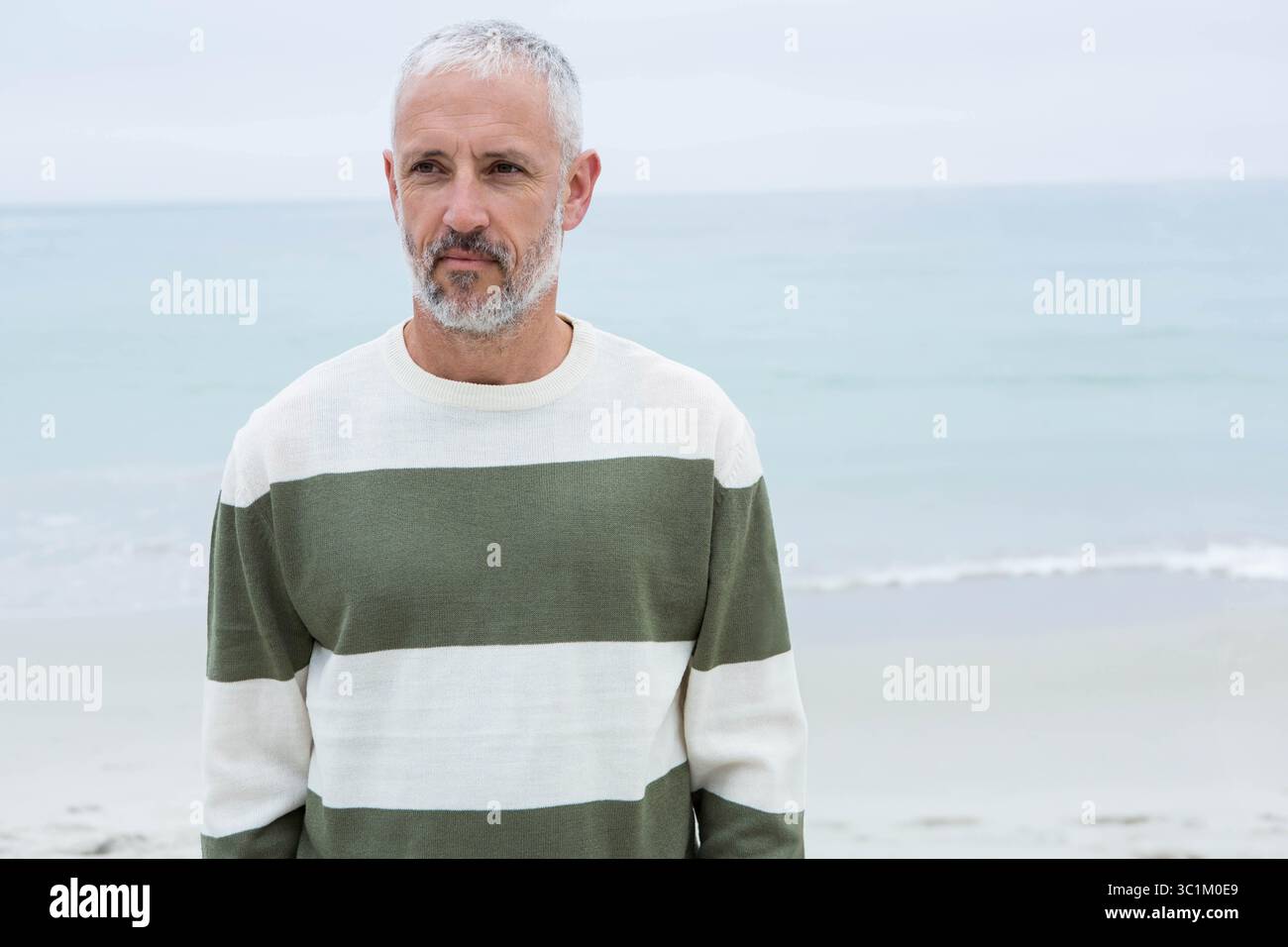 Homme senior debout sur le rivage de sable à la plage portant un pull à col ras du cou rayé vert olive Banque D'Images