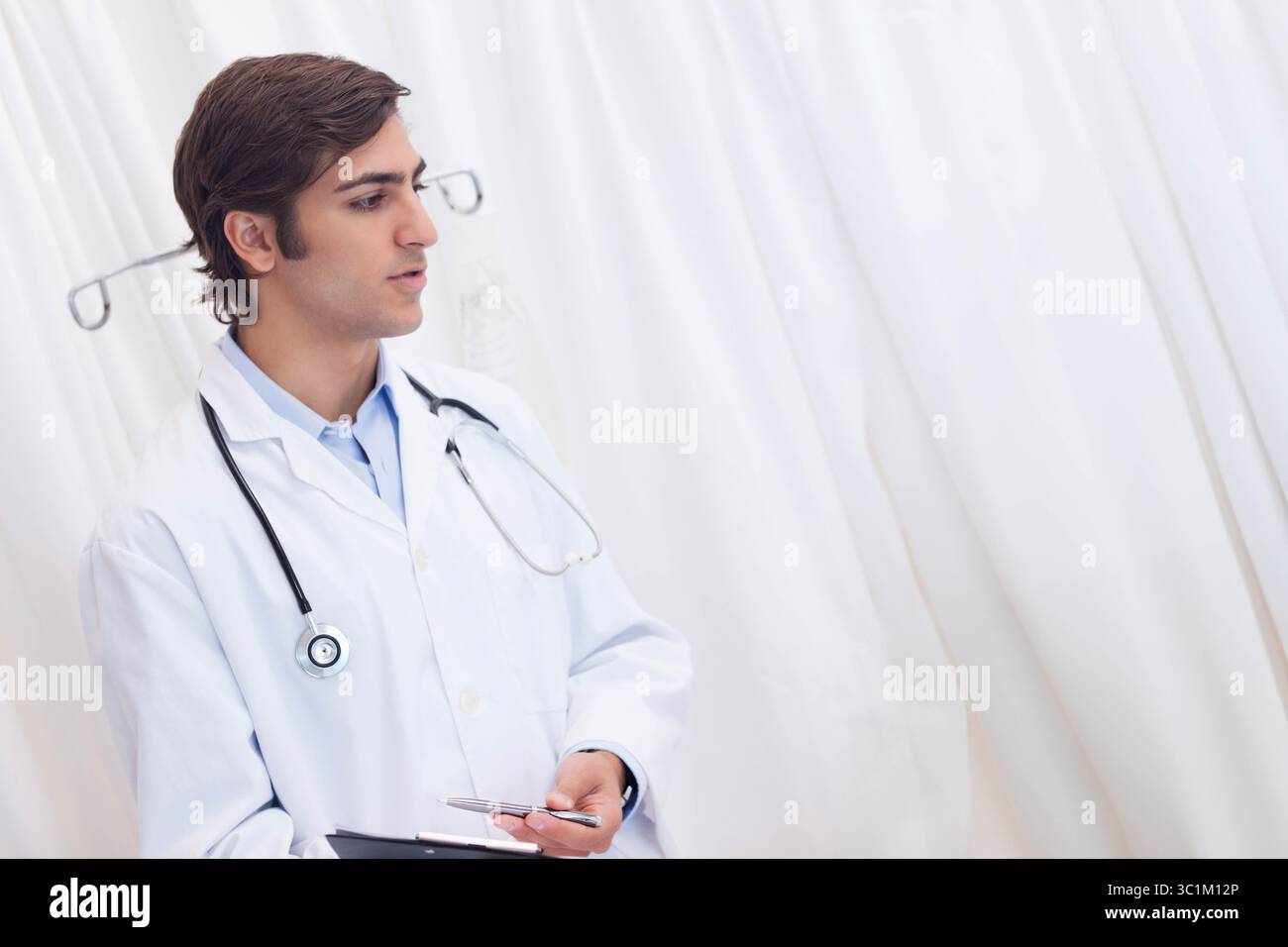 Médecin masculin debout à côté du rideau et du poteau IV en clinique portant un stéthoscope, tenant une presse-papiers Banque D'Images