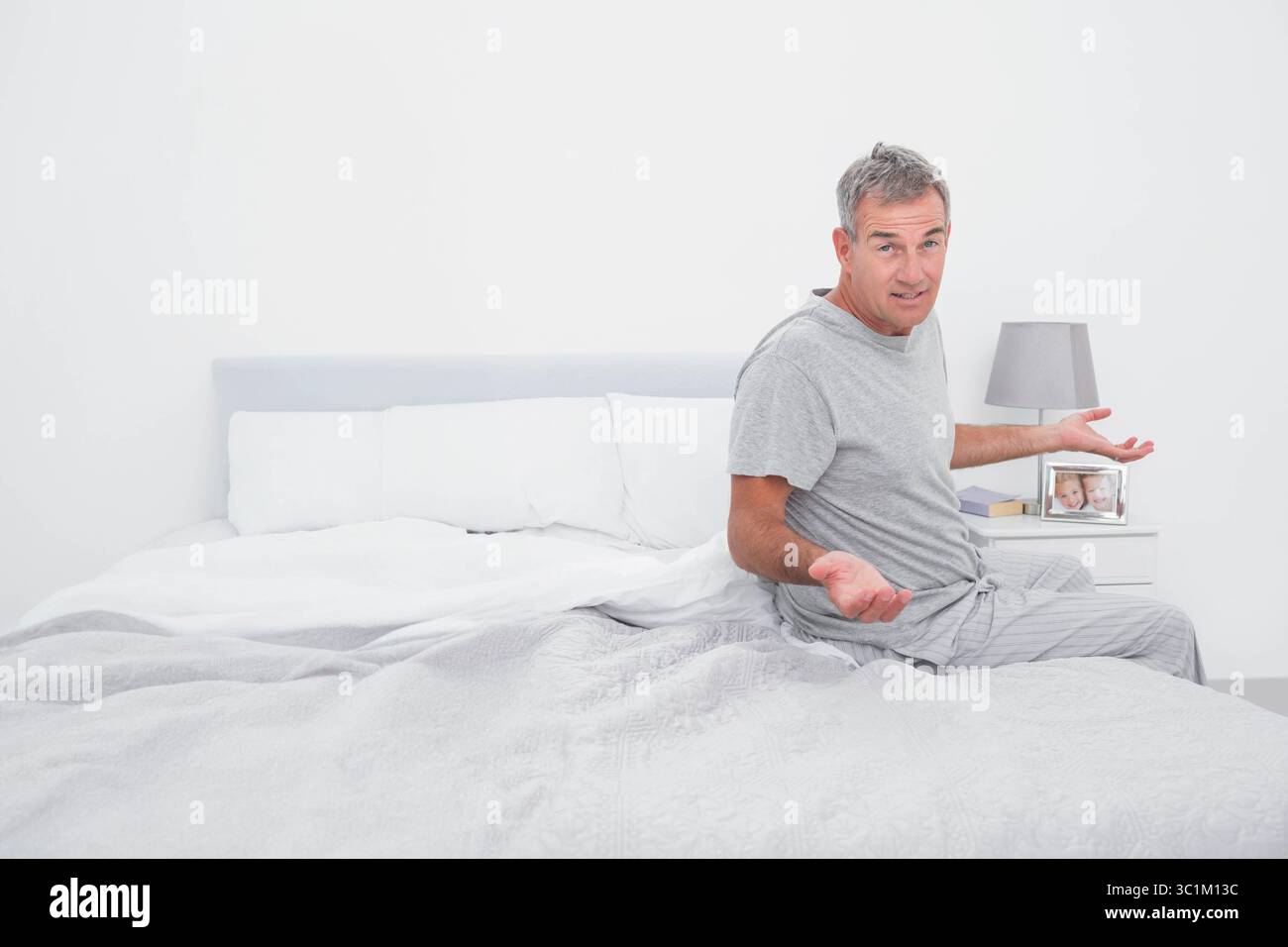 Homme senior haussant les paumes, assis sur le lit dans la chambre à coucher à côté de la table de chevet avec lampe, espace de copie Banque D'Images