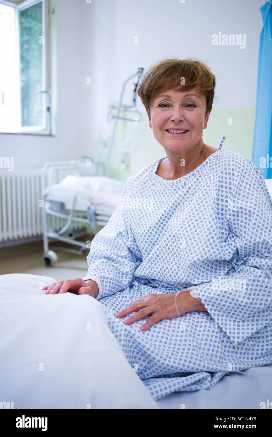 Femme âgée assise à l'hôpital sur le lit tenant la ligne IV en utilisant la barre de trapèze sur le lit portant la robe Banque D'Images