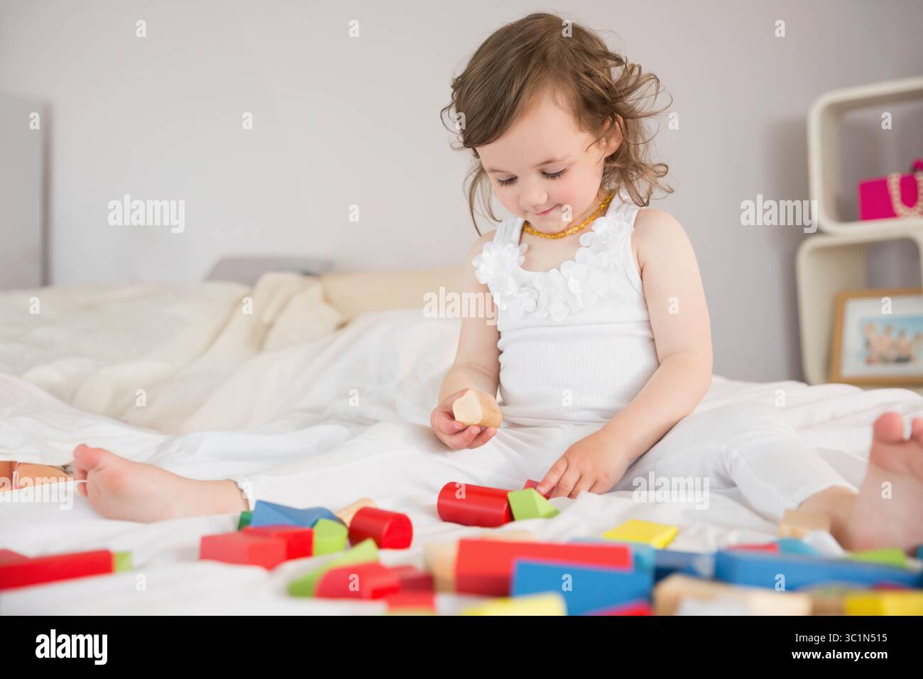 Jeune fille jouant avec des blocs de bois colorés sur des draps de lit blancs dans une chambre à coucher légèrement éclairée Banque D'Images