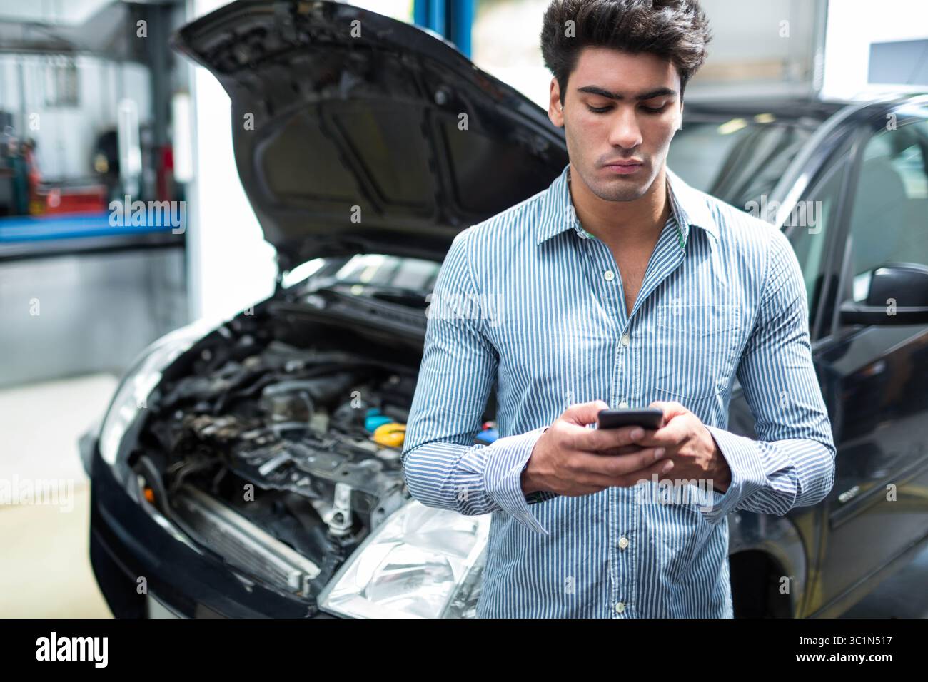 Homme portant chemise rayée debout dans un atelier de réparation automobile tenant smartphone près de la voiture avec le capot ouvert Banque D'Images