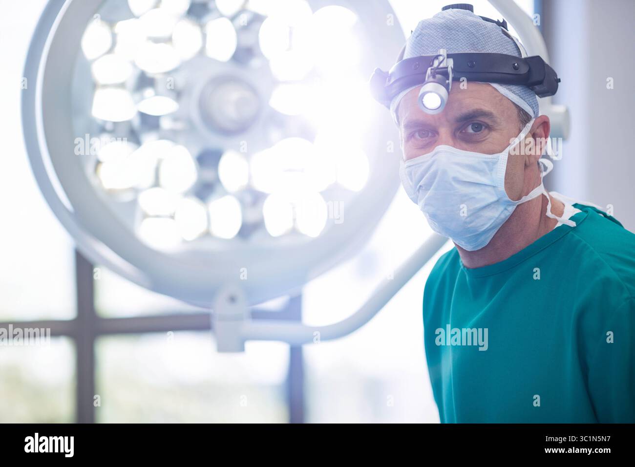 Chirurgien d'âge moyen debout sous la lumière LED, portant des gommages verts et une lampe frontale dans la salle d'opération Banque D'Images