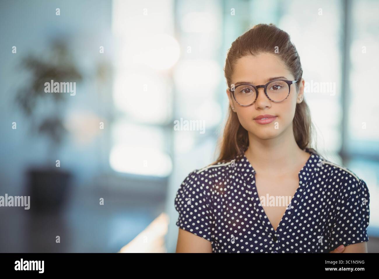 Femme portant des lunettes, chemisier à pois bleu marine debout par une plante en pot dans le hall du bureau, espace de copie Banque D'Images