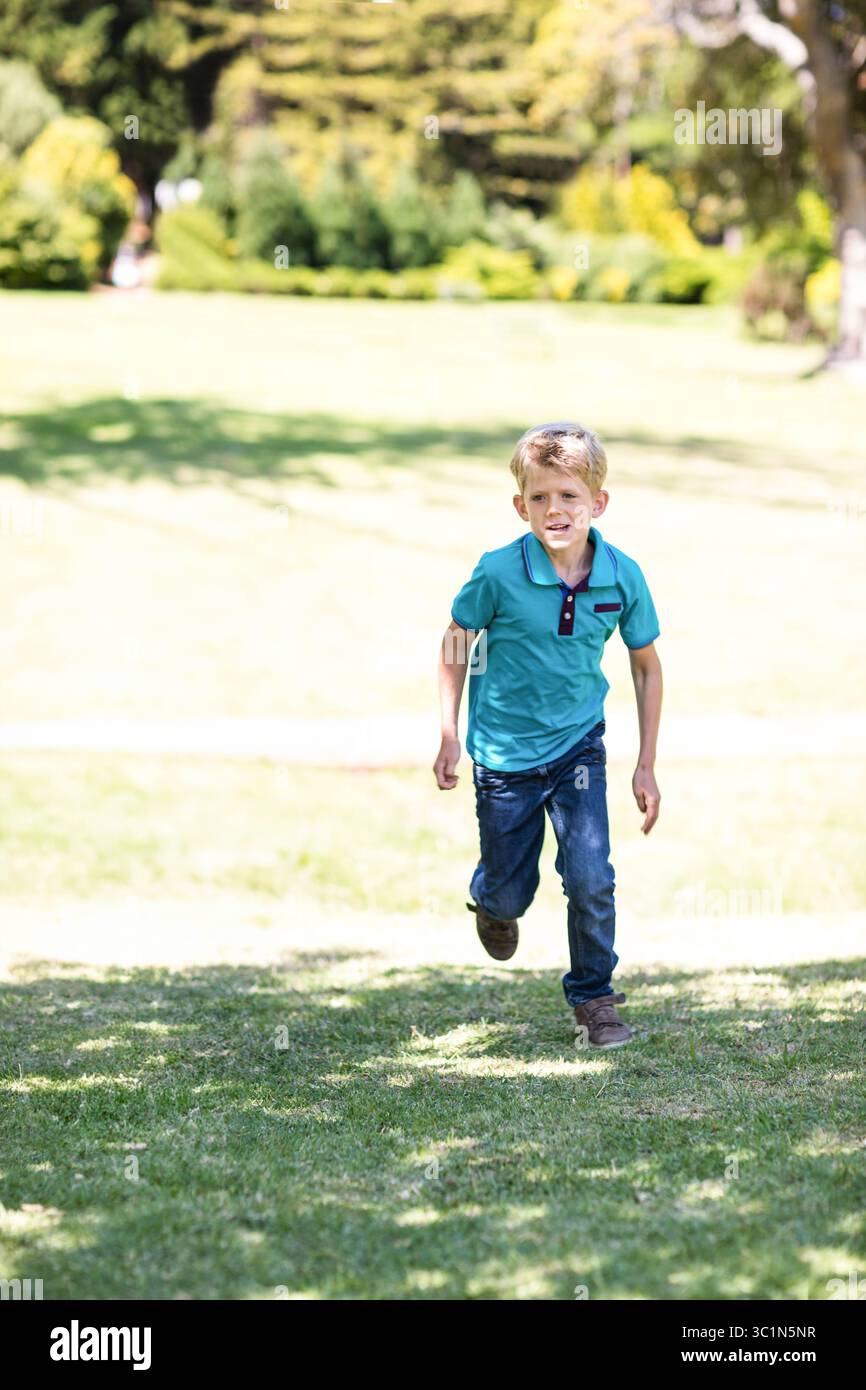 Garçon qui court à travers un terrain de parc herbeux ensoleillé portant un polo turquoise et un Jean foncé Banque D'Images