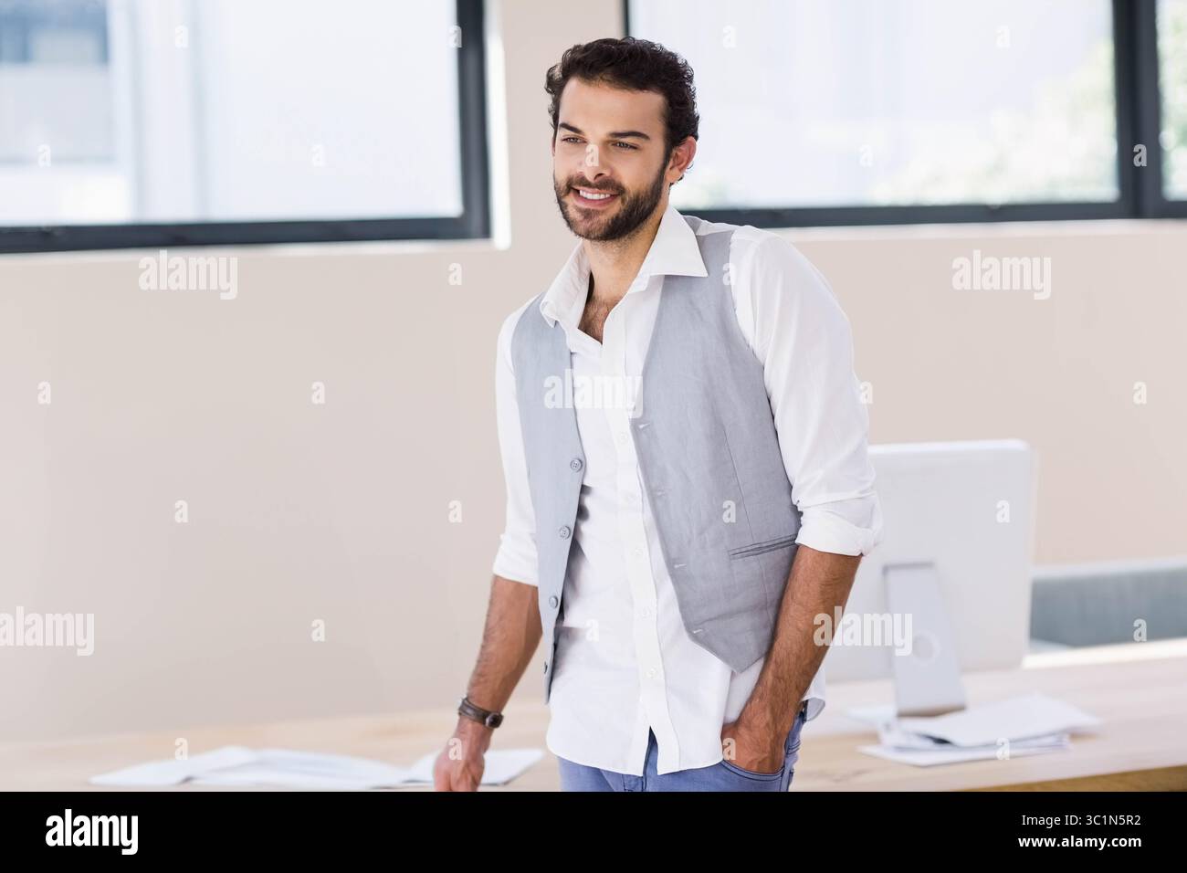 Homme souriant doucement tout en s'appuyant sur un bureau en bois dans un bureau moderne avec ordinateur et papiers dispersés Banque D'Images