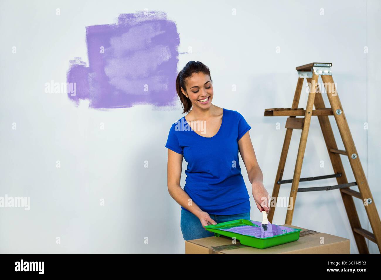 Femme peignant un mur blanc avec de la peinture violette à la maison à l'aide d'un pinceau et d'un escabeau en bois Banque D'Images