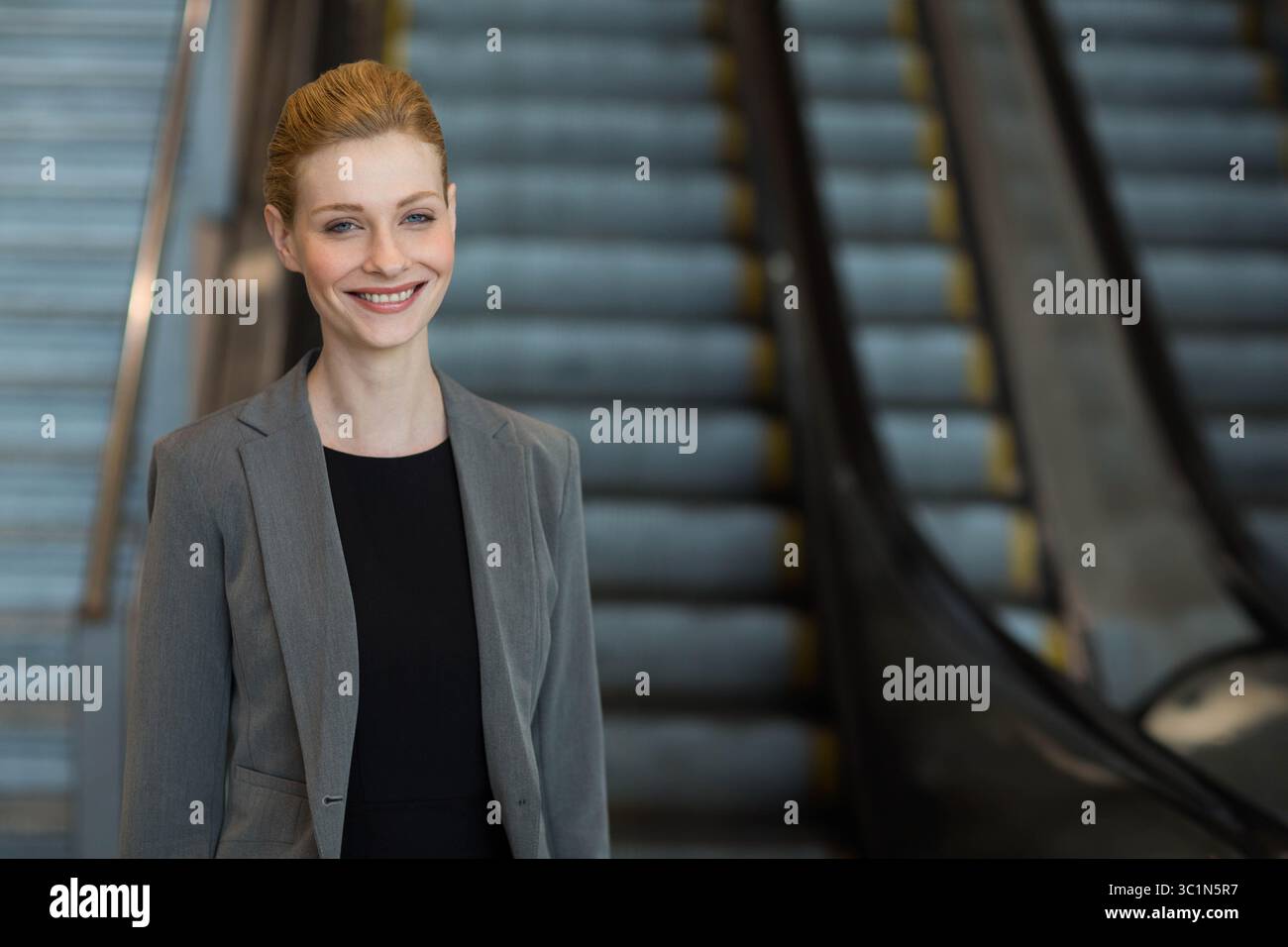 Femme d'affaires debout et souriante dans le hall près des escalators avec des mains courantes en métal, espace de copie Banque D'Images