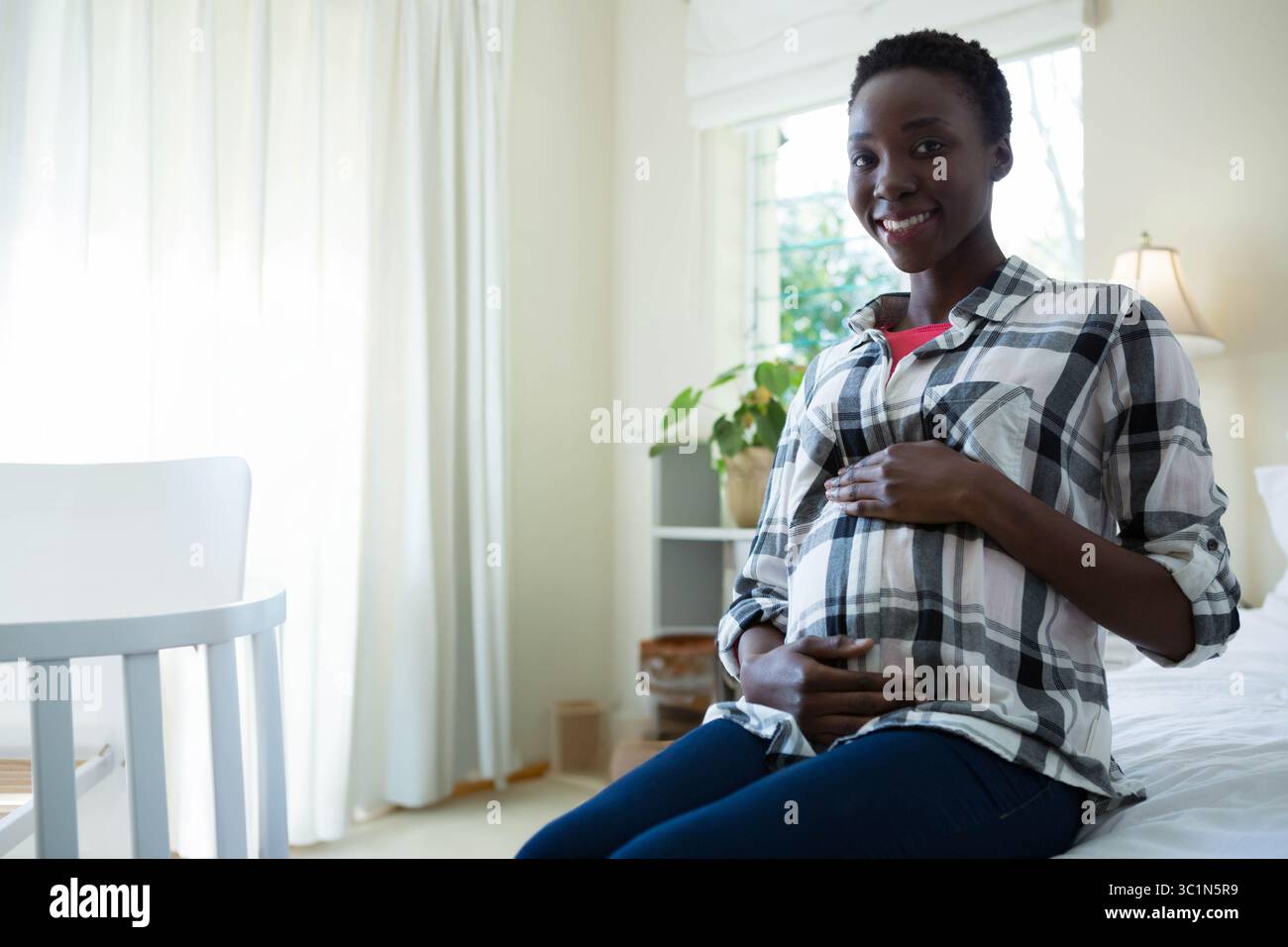 Femme afro-américaine tenant le ventre enceinte assis sur le lit dans la pépinière avec berceau blanc, espace de copie Banque D'Images
