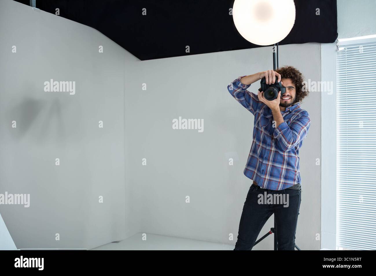 Photographe masculin tient un appareil photo reflex numérique et vise dans un studio d'angle blanc avec lumière Softbox Banque D'Images