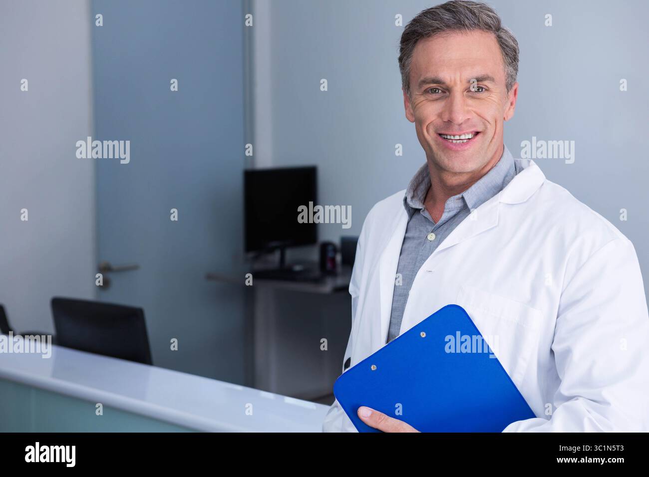 Homme portant une blouse de laboratoire debout près de l'écran de l'ordinateur à la réception tenant un presse-papiers bleu, espace de copie Banque D'Images