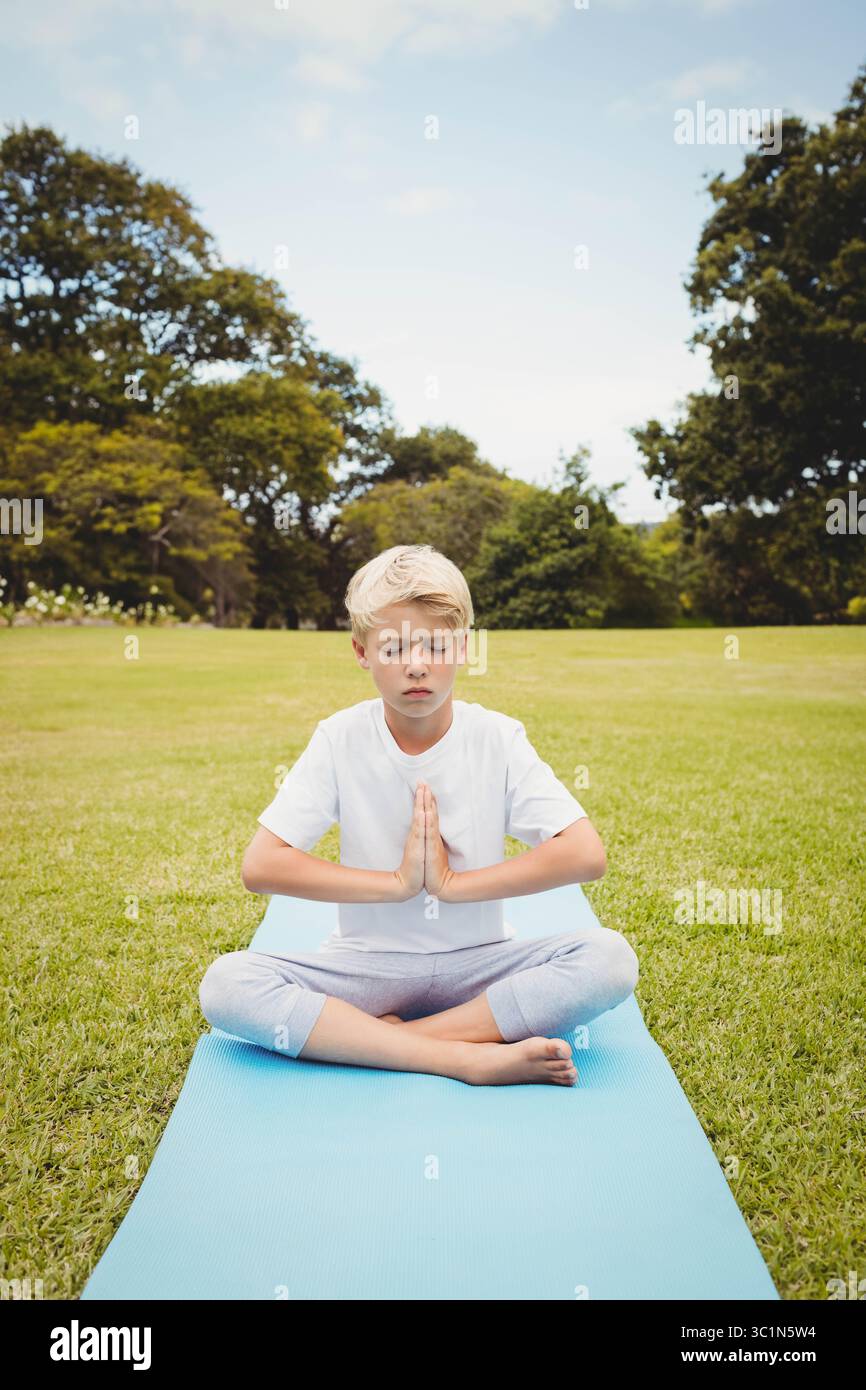 Garçon méditant les jambes croisées sur le tapis de yoga bleu vif dans le parc appuyant les mains à la poitrine Banque D'Images