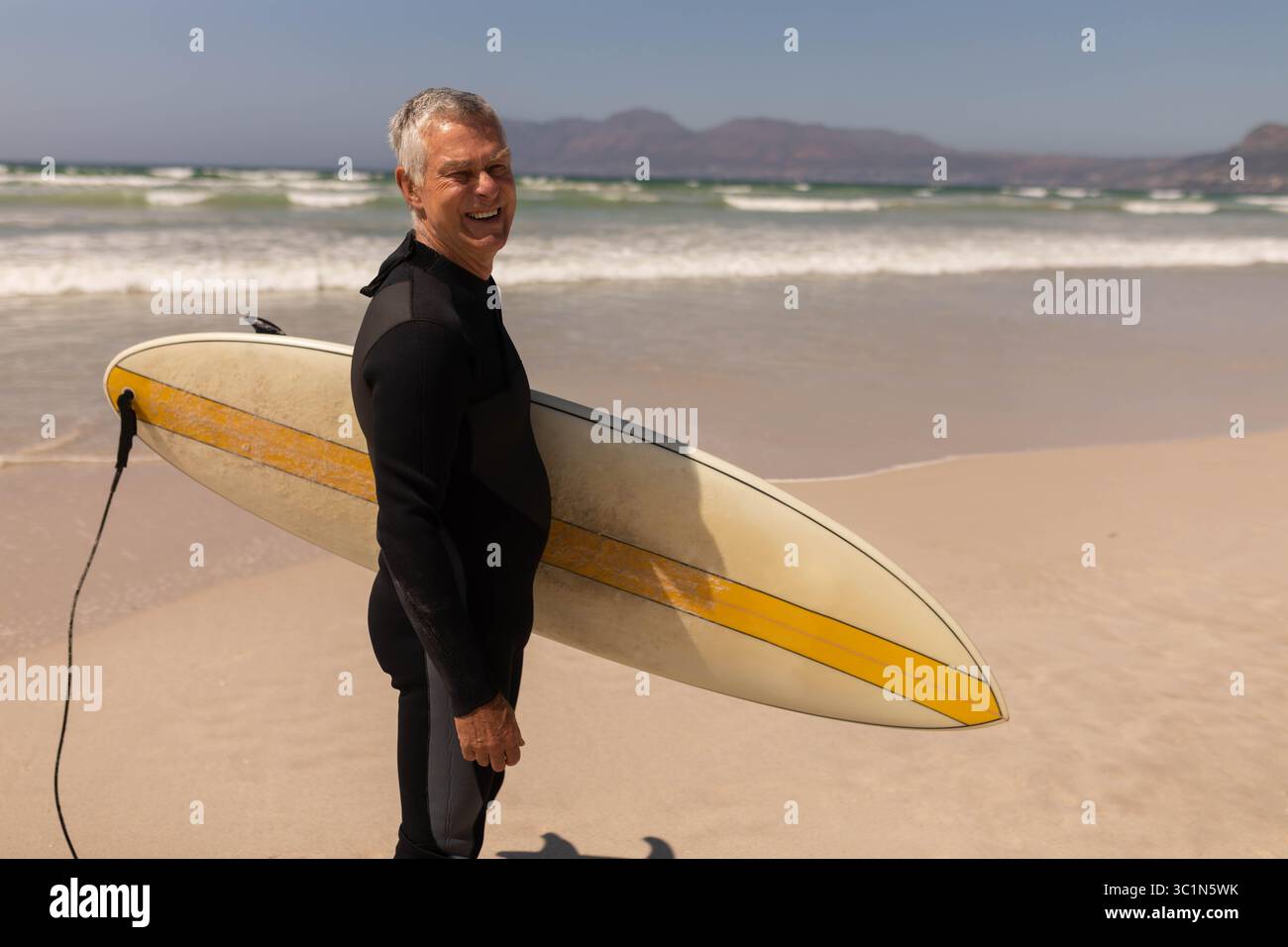 Surfeur masculin senior marchant le long de la plage portant une combinaison de plongée tenant une planche avec une bande jaune, espace copie Banque D'Images