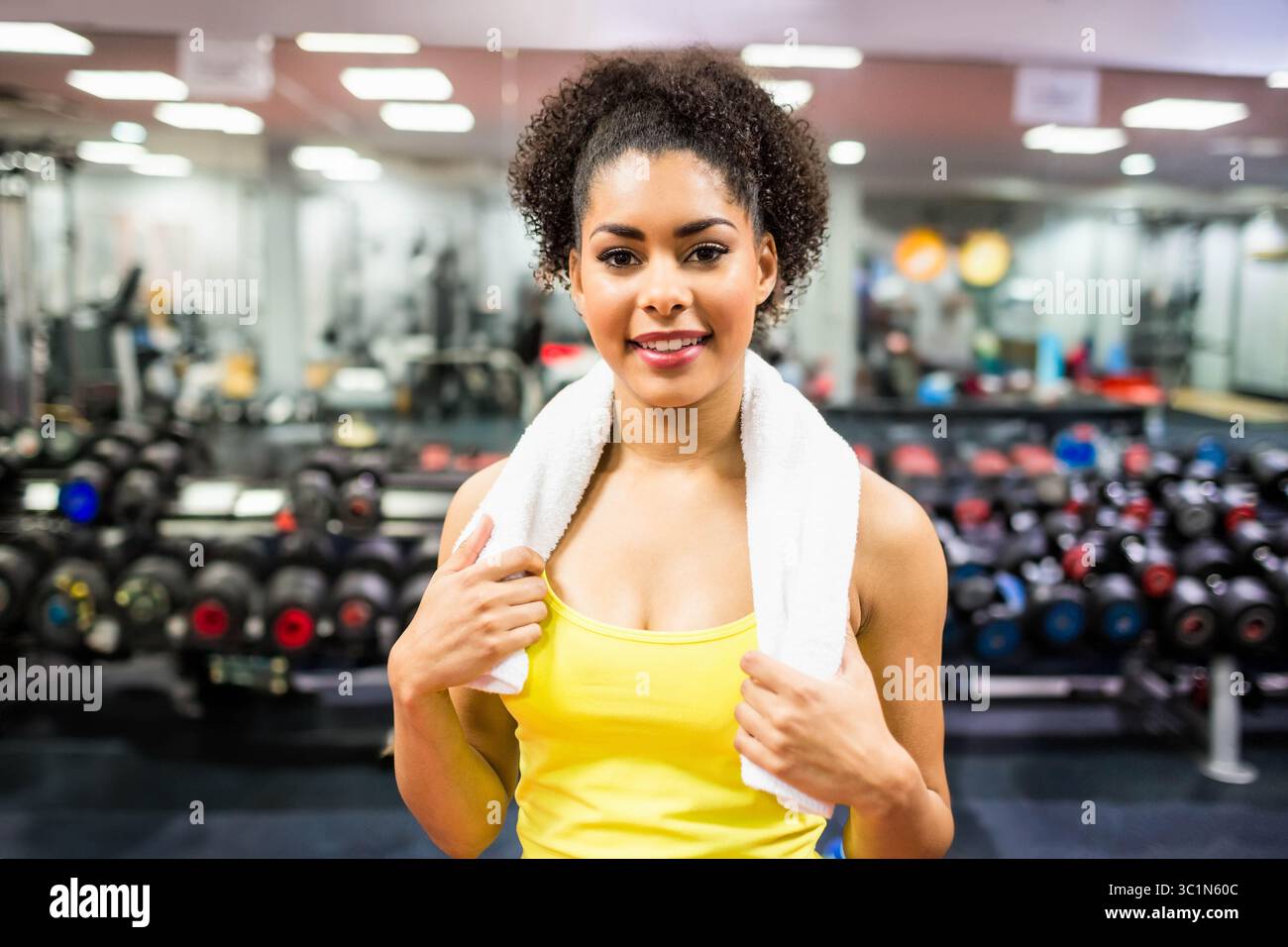 Femme afro-américaine portant un débardeur jaune et tenant une serviette blanche près du porte-haltères au gymnase Banque D'Images