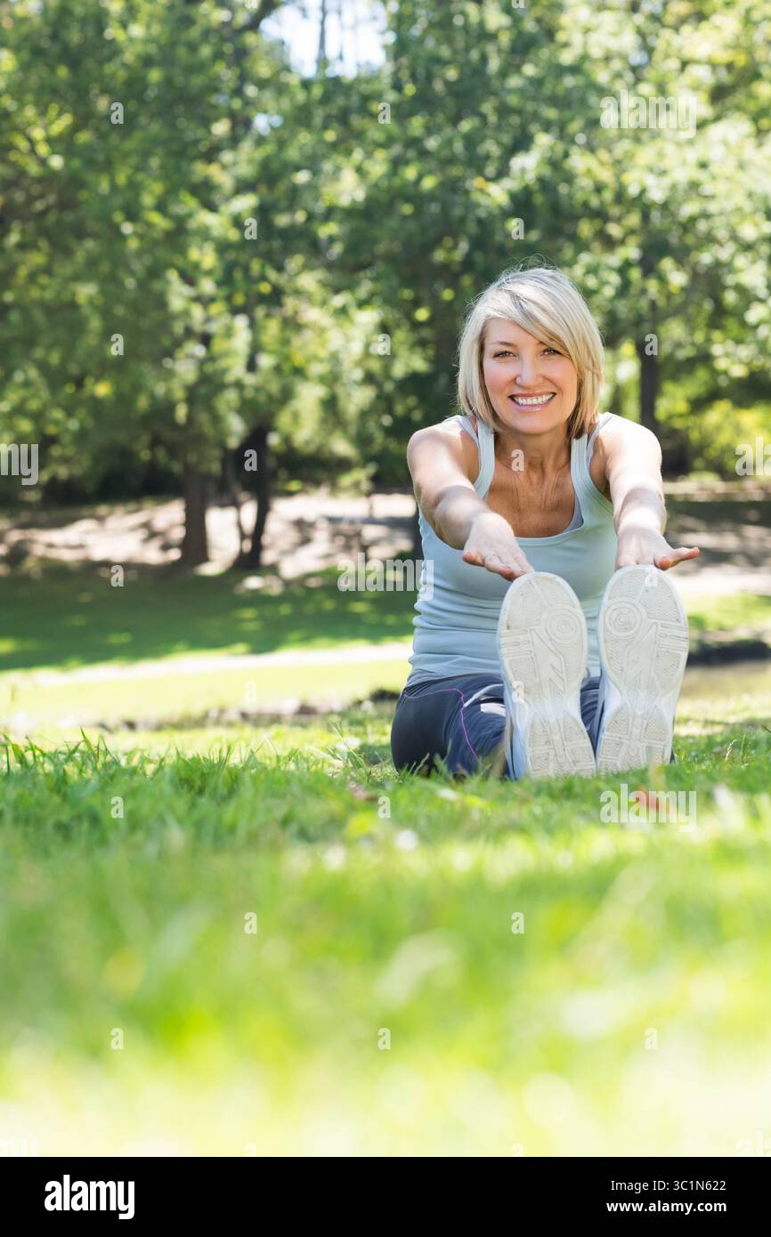 Femme d'âge moyen assise sur l'herbe près de l'étang dans le parc touchant les orteils tout en portant des chaussures athlétiques blanches Banque D'Images