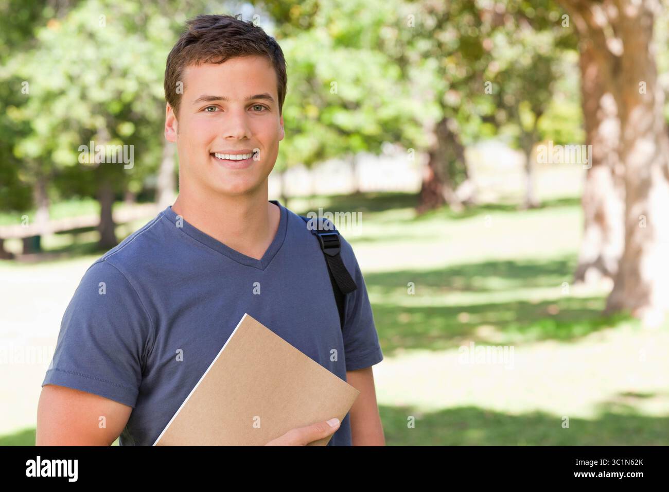 Étudiant masculin portant un T-shirt bleu sur la pelouse du campus, tenant un dossier et un sac à dos sous la lumière du soleil Banque D'Images