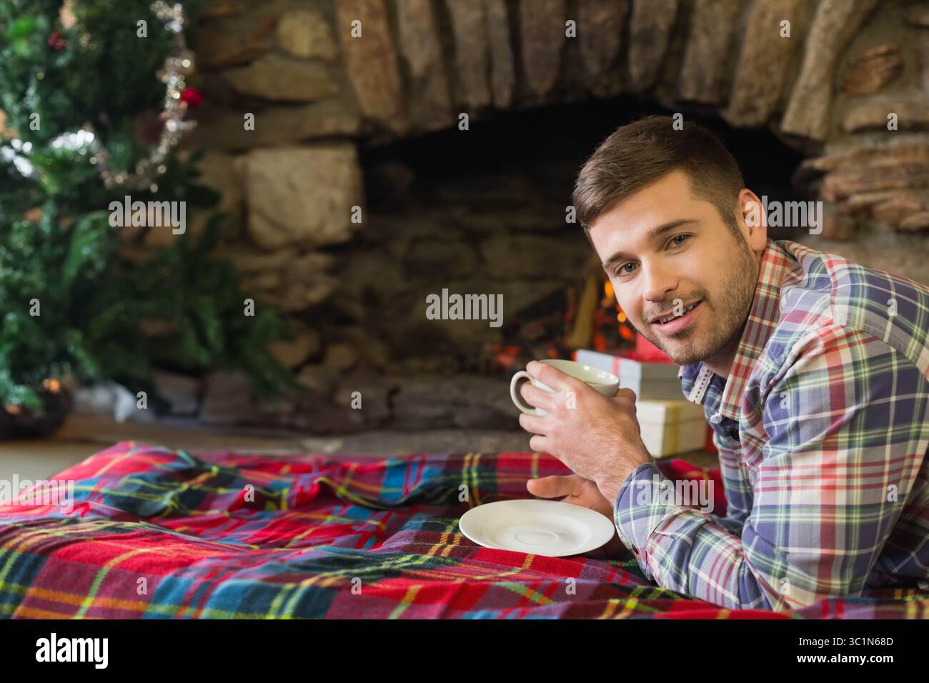 Homme incliné à la maison sur la couverture de tartan tenant la tasse par la cheminée, arbre de Noël, espace de copie Banque D'Images