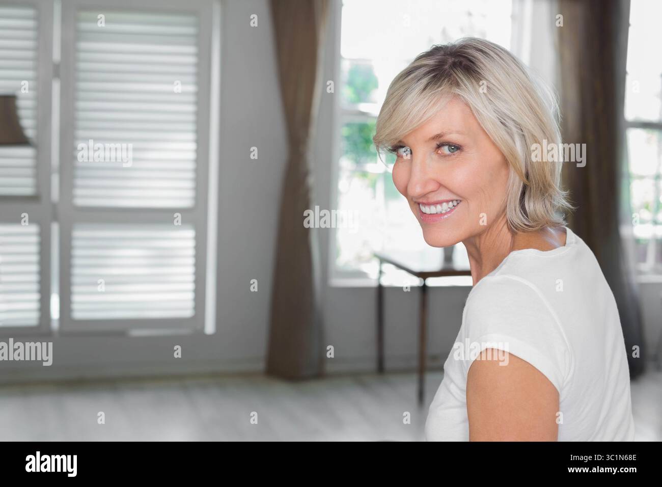Femme du milieu des années 40 souriant et regardant par-dessus l'épaule dans le salon par volets et table d'appoint Banque D'Images