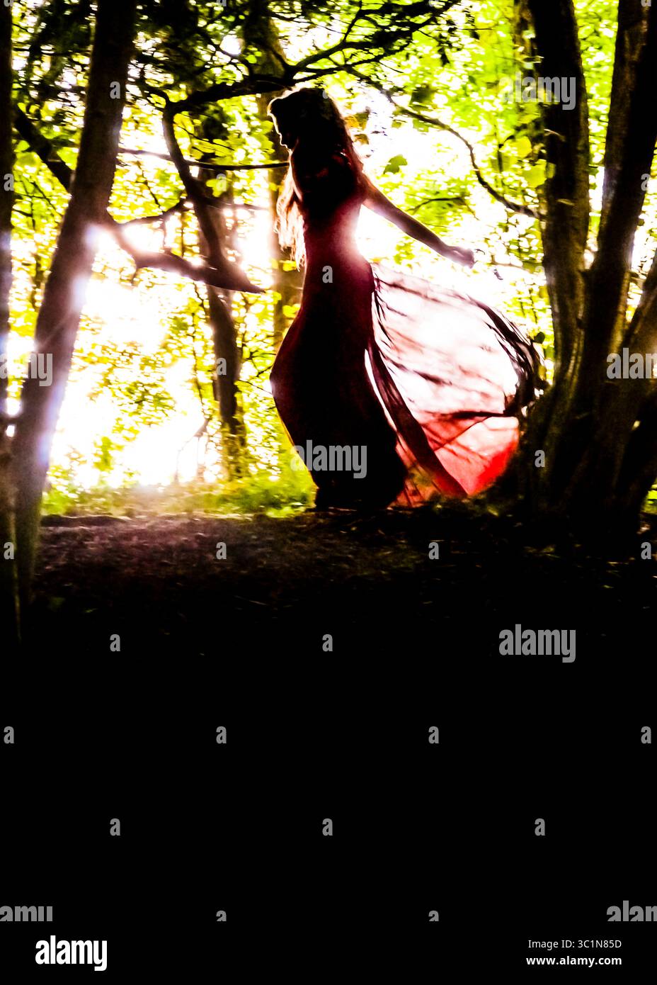 Une femme dans une robe rouge fluide se silhouette contre la lumière du soleil brillante filtrant à travers les arbres. Banque D'Images