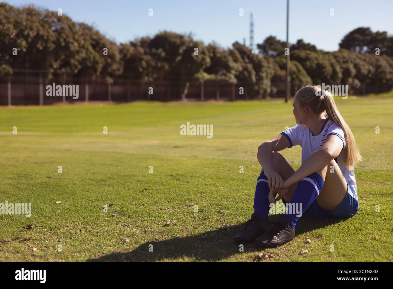 Joueur de football assis sur un terrain en herbe au parc, portant un maillot blanc, un short bleu et des crampons noirs Banque D'Images