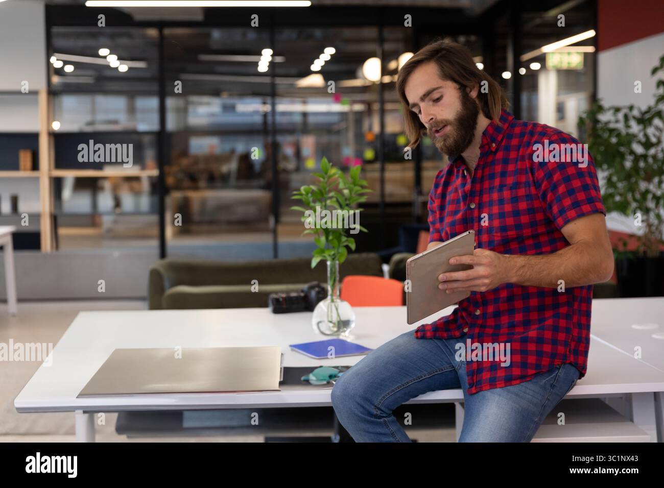 Homme perché sur le bureau étudiant tablette au milieu de l'ordinateur portable et vase avec branche verte dans le bureau ouvert Banque D'Images