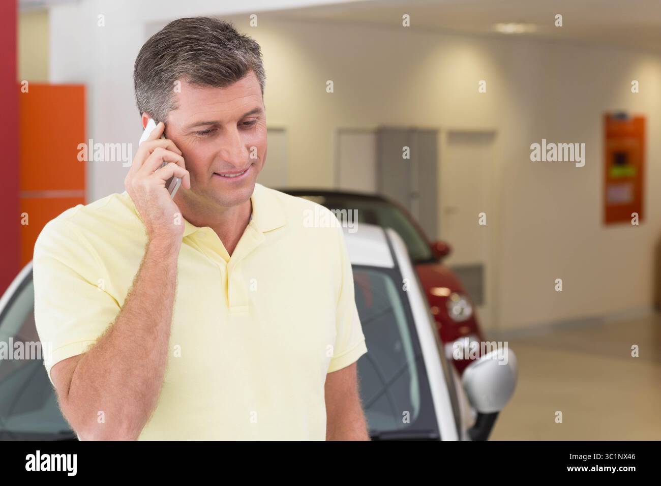 Homme senior appelant sur smartphone et inspectant la voiture bicolore avec panneau rouge dans la salle d'exposition de la concession Banque D'Images