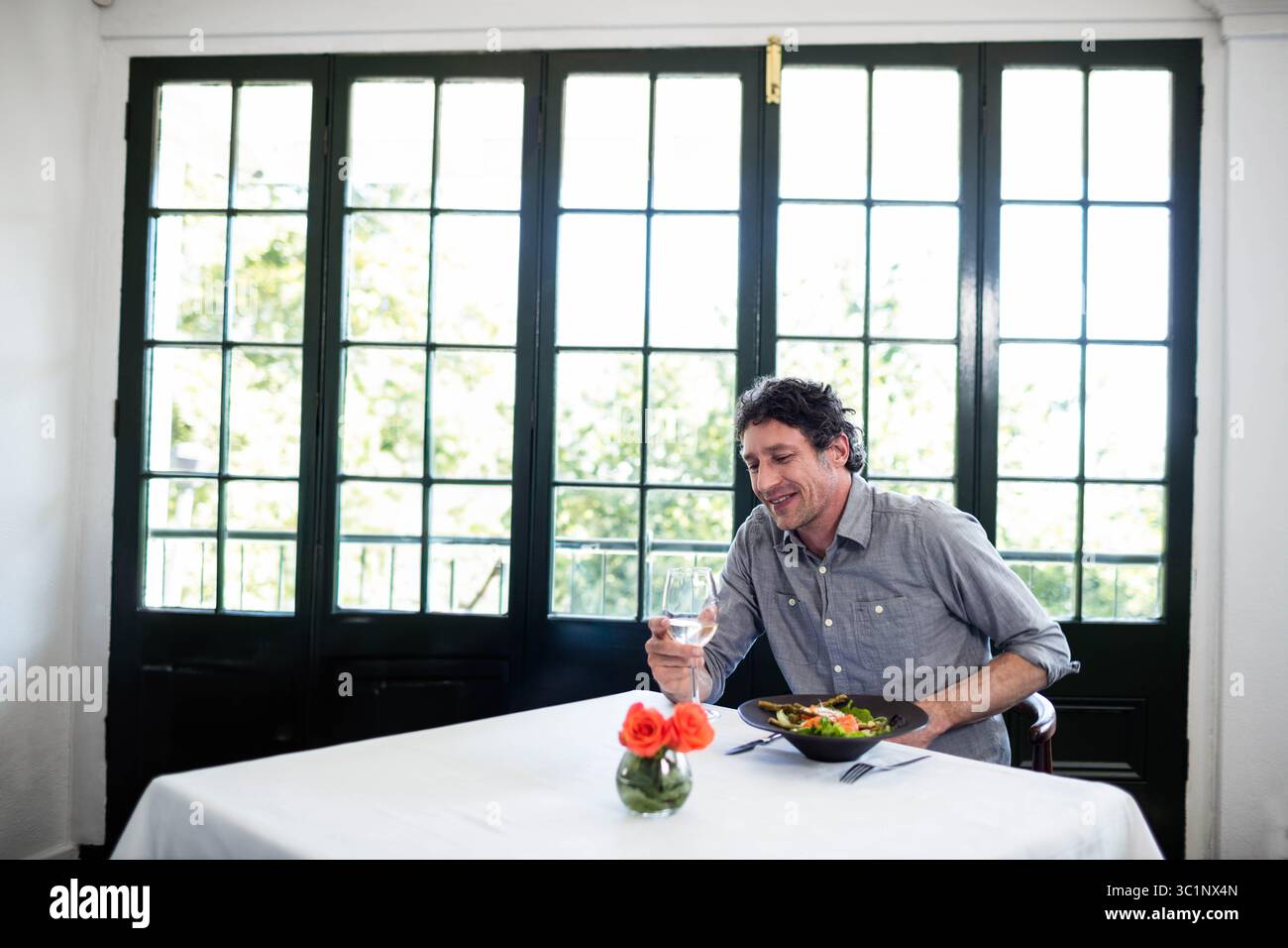 Verre à vin repose sur une nappe blanche dans la salle à manger, avec des roses orange par des fenêtres de grille Banque D'Images